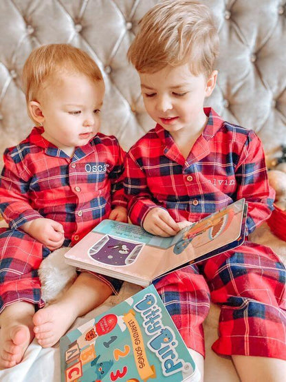 Two young children in matching red plaid pajamas sit on a bed, reading Ditty Bird UK’s Learning Songs together. The older child holds the interactive sound book open while the younger looks with fascination at its colorful educational pages.