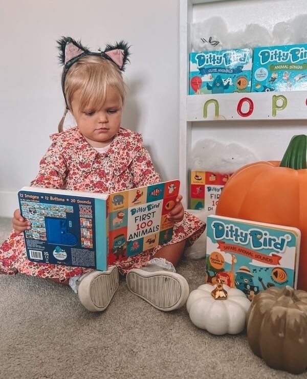 A young child wearing cat ear headbands and a floral dress sits on the floor, reading. Around her are Ditty Bird UK Safari Animal Sounds toys—an educational, musical gift for toddlers 1-3—plus a large orange pumpkin and two small decorative pumpkins.