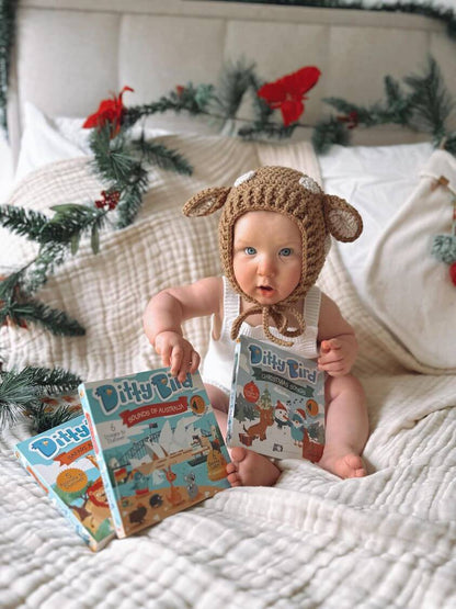 A baby in a knitted animal hat sits on a bed with holiday greenery, holding Ditty Bird UK's "Sounds of Australia" interactive sound book, with two more early learning Ditty Bird books nearby on a white blanket.