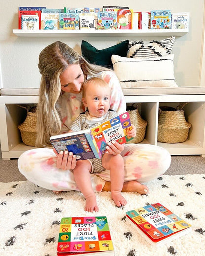 A woman sits on the floor with a smiling baby in her lap as they enjoy the Ditty Bird UK 100 Places Sound Book. Colorful baby toys for 6-12 months are scattered on the rug, with a shelf of books visible in the background.